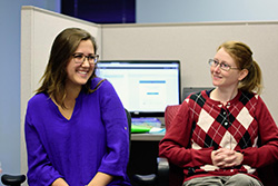 Two women smiling and sitting in front of a computer screen in an office cubicle