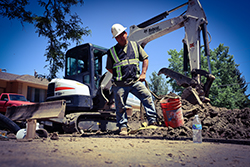 Male construction worker standing near a backhoe inspecting a digging project in the street